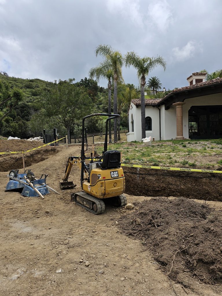 Compact excavator cutting a deep irrigation trench at a Spanish-style hillside estate