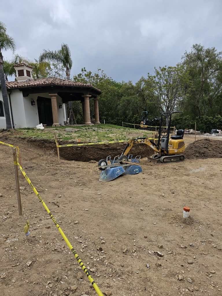 Mini-excavator digging new irrigation and landscape lines beside a Spanish-style pool pavilion